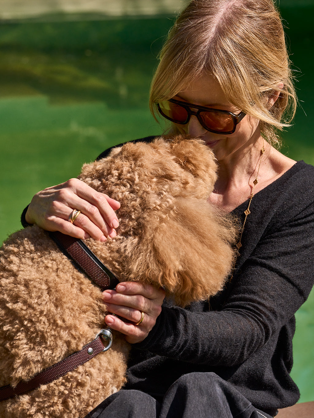 Woman cuddling a curly dog wearing a premium brown leather collar beside a lake.