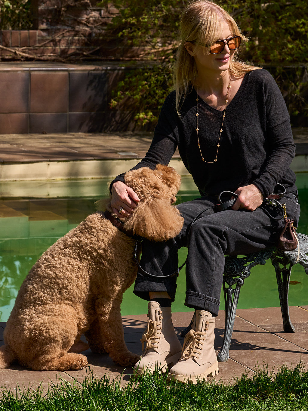Woman in black outfit sitting poolside with apricot poodle on leather leash, wearing tan boots