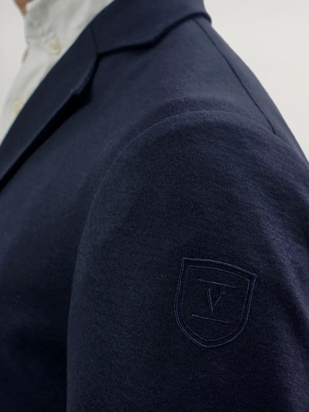 A close-up of a navy blue equestrian jacket shoulder featuring a tonal embroidered shield logo with the letter "V".
