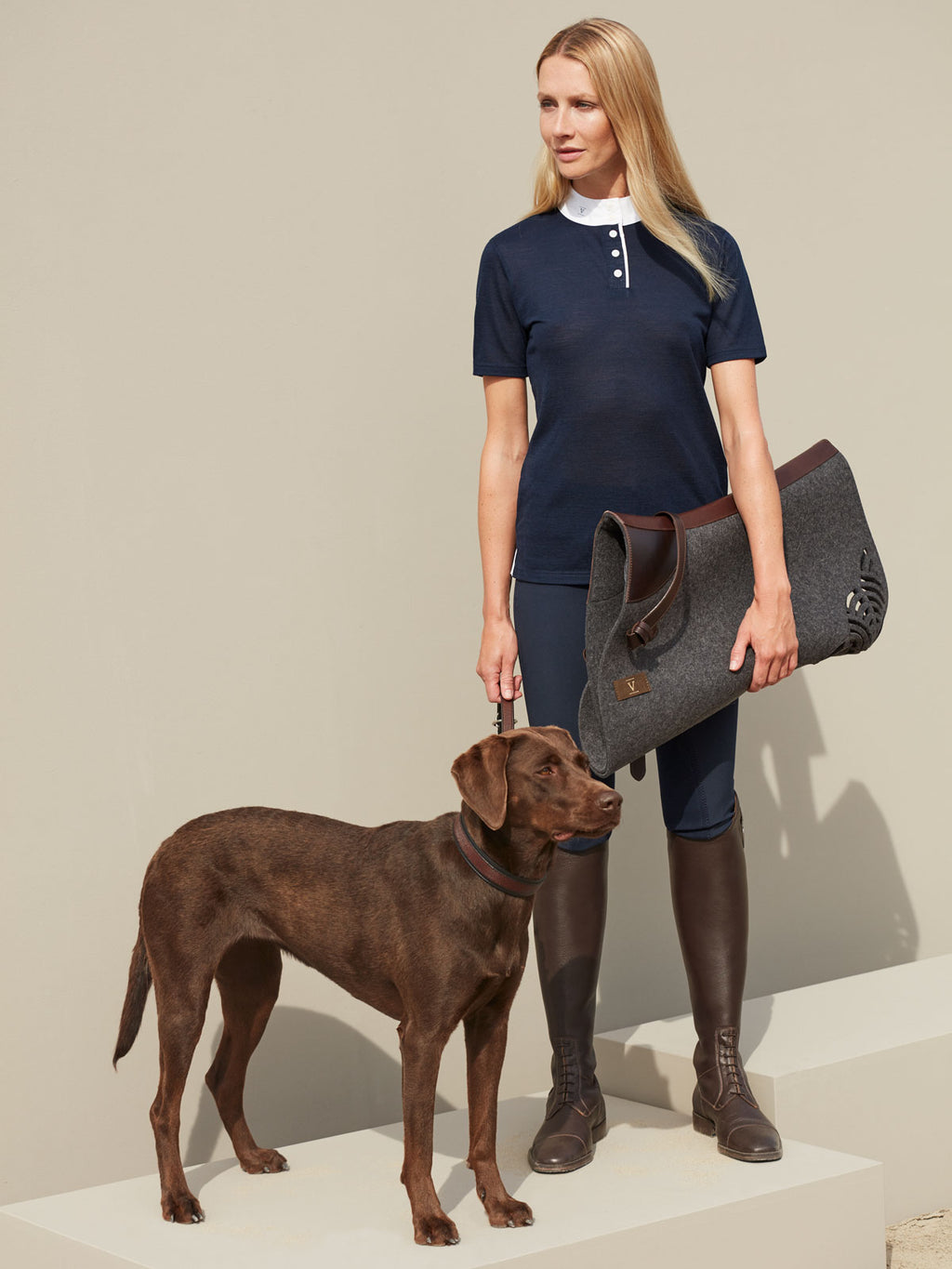 A female rider in navy equestrian apparel holding a grey felt saddle pad and standing with a chocolate Labrador.