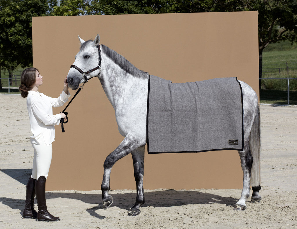 Woman in equestrian attire with a dapple grey horse wearing a grey wool blanket in an outdoor arena.