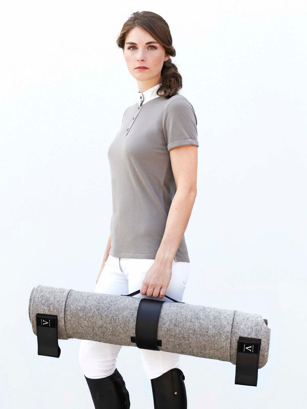 Woman in equestrian riding attire carrying rolled grey wool felt Manifattura Valor dog bed with black leather straps.